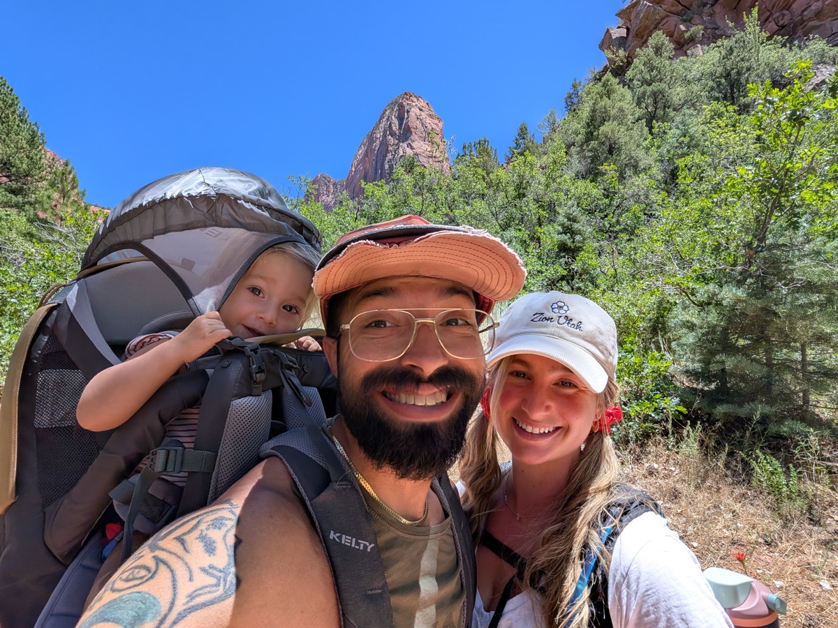 Joe Lucky smiling on a trail with his wife and their daughter riding in a backpack carrier, a red rock peak rising behind them in Zion, Utah.